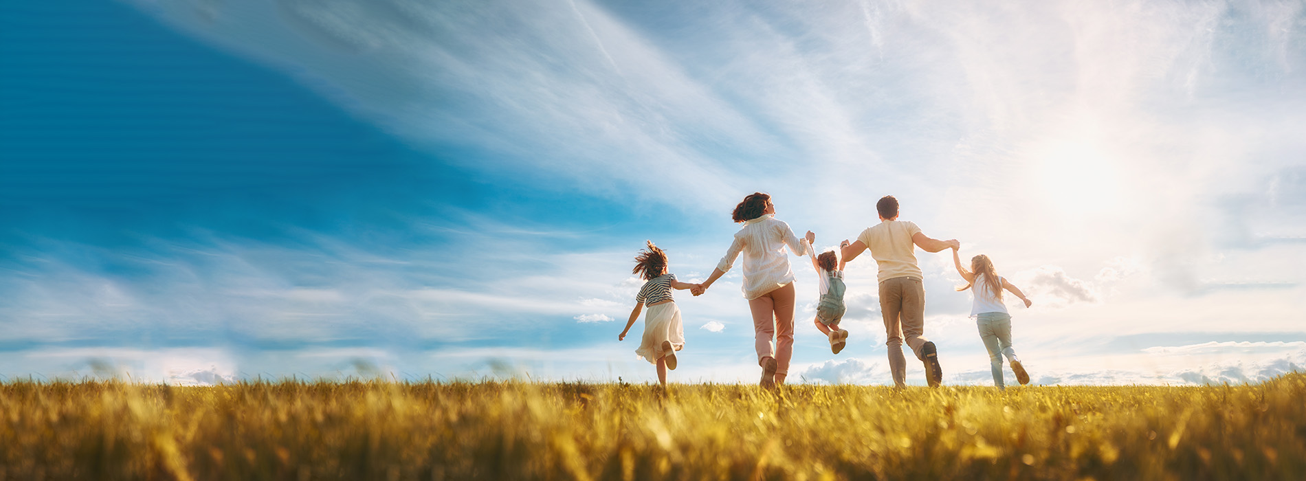 The image depicts a family of four, including two adults and two children, standing on a grassy hillside with their arms outstretched towards the sky, embracing each other in a moment of happiness against a backdrop of a clear blue sky.